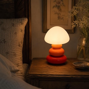 May include: A mushroom-shaped table lamp featuring a glowing white glass dome shade atop a stacked base composed of three rounded, glossy ceramic sections in graduated shades of orange, ranging from a light orange to a deep, vibrant red-orange.