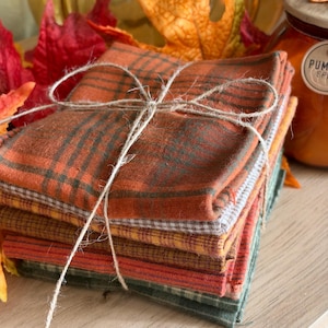 May include: A stack of four folded fabric squares tied together with thin twine. The top square features an orange and dark green plaid pattern, followed by a grey gingham square, a yellow plaid square, and a dark green plaid square.