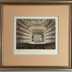 May include: Framed antique print depicting an opera house interior. The artwork shows a detailed view of the theater, with multiple levels of seating filled with figures. The stage is visible, and the print is framed in a gold-colored frame.
