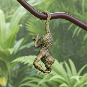 Op de afbeelding: Een bronzen aapfiguur die ondersteboven aan een bruine tak hangt. De aap heeft een gedetailleerde vachttextuur en staat tegen een achtergrond van weelderig groen gebladerte, wat een junglethema creëert.