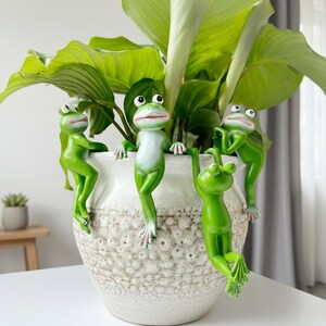 May include: A white ceramic planter with a textured surface, filled with green leafy plants. Four green frog figurines are attached to the planter, appearing to climb and hang from the rim. The frogs have white bellies and are in various poses.
