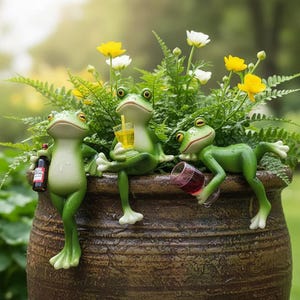 Puede incluir: Tres figuras de ranas verdes están posadas en una maceta marrón, cada una sosteniendo una bebida. Una rana sostiene una botella, otra una bebida amarilla con una pajita y la tercera una copa de vino tinto. La maceta está llena de plantas verdes y flores amarillas.