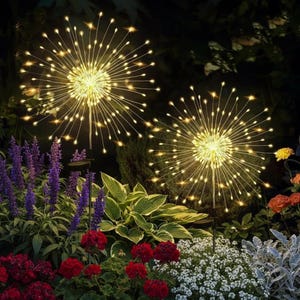 Può includere: Due luci da giardino a energia solare con un design a forma di fuoco d'artificio. Ogni luce ha una sfera centrale con luci LED bianche calde radianti. Le luci sono ambientate in un giardino con fiori e verde colorati.