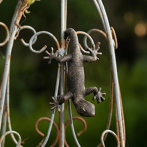 Puede incluir: Una escultura de lagarto de metal color bronce sujeta a un arco de jardín decorativo de metal blanco desgastado. El lagarto tiene una textura detallada y está colocado boca abajo. El fondo es verde borroso.