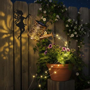 Puede incluir: Una regadera decorativa con energía solar y silueta de hada. La regadera de color bronce tiene un diseño floral y emite un brillo cálido, con luces de cadena que caen en una maceta de terracota llena de vegetación y flores moradas. El conjunto se encuentra frente a una valla de madera.