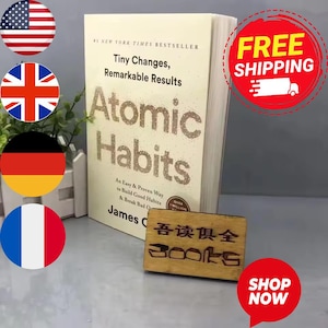 May include: A hardcover book titled "Atomic Habits" by James Clear. The book has a beige cover with the title in large, glittery letters. The book is a New York Times bestseller and promises to help build good habits. A small wooden sign is in front of the book.