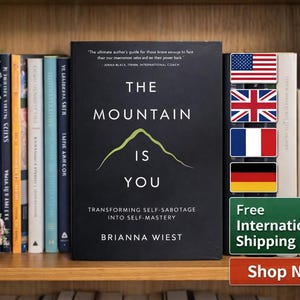 May include: A hardcover book titled "The Mountain Is You" by Brianna Wiest, with a green mountain graphic. The book is on a shelf with other books, flowers, and a small succulent. The image includes flags and a "Shop Now" button.