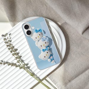 May include: A light blue smartphone case featuring three white Hello Kitty character figures arranged vertically. Each figure has a small blue bow on its head. The case has a clear border and cutouts for the dual camera lenses on the back.