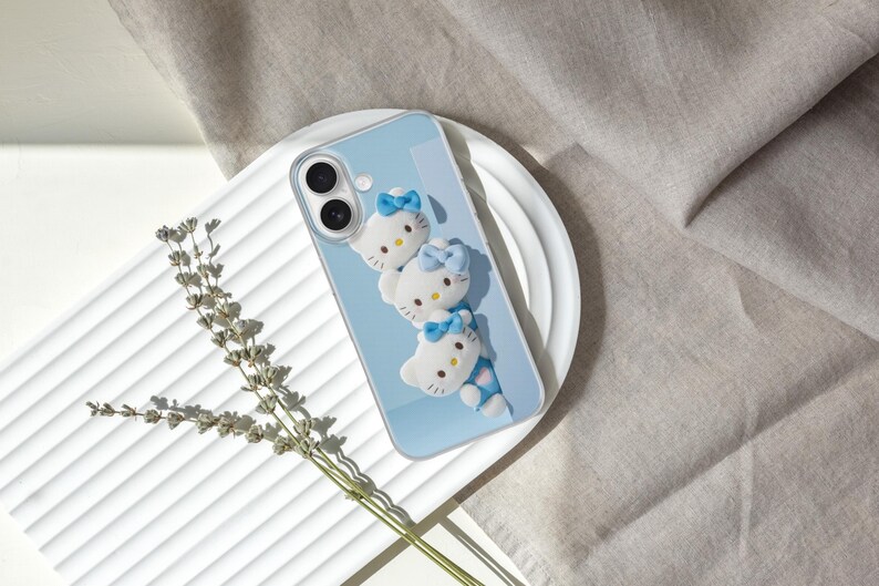 May include: A light blue smartphone case featuring three white Hello Kitty character figures arranged vertically. Each figure has a small blue bow on its head. The case has a clear border and cutouts for the dual camera lenses on the back.