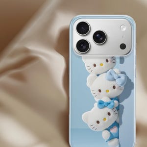 May include: A clear smartphone case featuring a light blue background with three stacked white Hello Kitty figures. Each figure has a yellow nose, black eyes, and whiskers. Two of the figures wear light blue bows, and one wears a blue outfit.