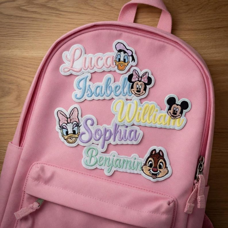 May include: A pink backpack with embroidered patches featuring Disney characters and names like "Luca," "Isabel," and "William." The patches include images of Donald Duck, Minnie Mouse, Daisy Duck, Mickey Mouse, and Chip.