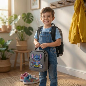 May include: A gray lunch bag featuring a blue alien character holding a pencil and books with the name Noah printed below. The bag has a black handle and zipper closure, designed for carrying food items in a rectangular, insulated soft-sided container.