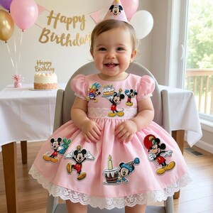 May include: A pink dress with Mickey Mouse graphics and white lace trim. The dress features short sleeves and is worn by a child. A party hat with Mickey Mouse is on the child's head. Balloons and a banner that says "Happy Birthday" are in the background.