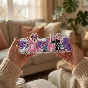 May include: A custom embroidered name patch spelling SOPHIA in a playful font. The letters feature a color palette of purple, pink, and white, decorated with cartoon character illustrations of Minnie Mouse and Daisy Duck, along with a pink polka dot bow.