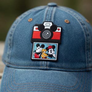 Puede incluir: Gorra de b&eacute;isbol de mezclilla azul con un parche colorido que presenta un dise&ntilde;o de c&aacute;mara roja y negra y una imagen de Mickey Mouse, Pluto y otro Mickey Mouse. La gorra tiene una visera curva y un bot&oacute;n en la parte superior.