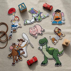K&ouml;nnte beinhalten: Eine Sammlung von gestickten Aufn&auml;hern mit Toy Story-Figuren, darunter ein Cowboy, ein Cowgirl, ein Weltraum-Ranger, eine kartoffelf&ouml;rmige Figur, ein Schwein, ein Dinosaurier, ein Federhund sowie ein Holzklotz mit dem Buchstaben A und zwei Spielzeugautos.