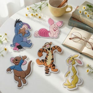 May include: Five embroidered fabric patches featuring cartoon characters: a blue donkey holding a bear, a pink piglet, a brown kangaroo, an orange and black striped tiger, and a yellow rabbit. The patches are arranged on a white surface.