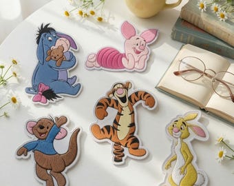 May include: Five embroidered fabric patches featuring cartoon characters: a blue donkey holding a bear, a pink piglet, a brown kangaroo, an orange and black striped tiger, and a yellow rabbit. The patches are arranged on a white surface.
