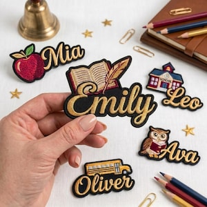 Puede incluir: Parches bordados con nombres y diseños escolares, incluyendo una manzana, un libro, una escuela, un búho y un autobús escolar. Los parches presentan letras doradas sobre un fondo negro. Una mano sostiene el parche "Emily".