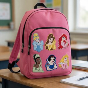 May include: Pink backpack featuring embroidered patches of Disney princesses. The backpack has a black top handle, black zippers, and adjustable shoulder straps. The patches include Cinderella, Belle, Ariel, Tiana, Snow White, and Aurora.