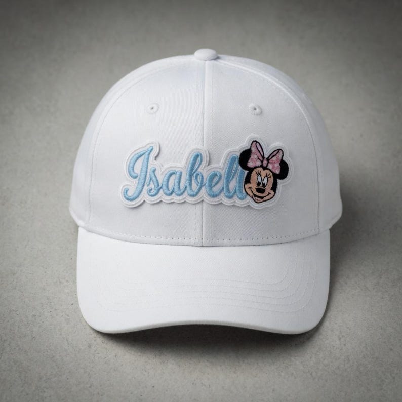 May include: White baseball cap featuring the name "Isabell" in light blue script with a Minnie Mouse patch. The Minnie Mouse patch has a pink bow. The cap is a classic six-panel design with a curved brim.