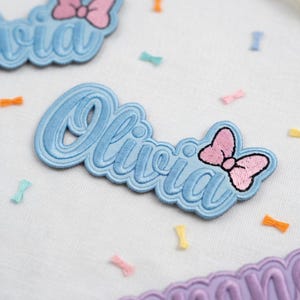 May include: Embroidered patch in light blue with the name "Olivia" in a cursive font. A pink bow is on the right side. The patch is surrounded by small, colorful bow-shaped confetti.
