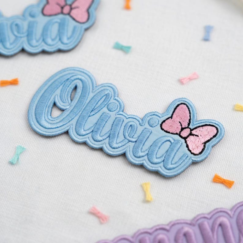 May include: Embroidered patch in light blue with the name "Olivia" in a cursive font. A pink bow is on the right side. The patch is surrounded by small, colorful bow-shaped confetti.