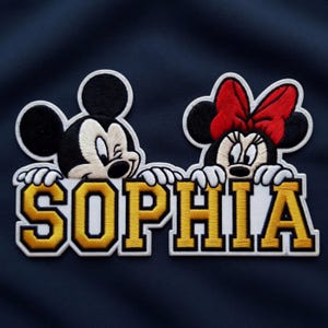 Puede incluir: Parche bordado con los personajes de Mickey y Minnie Mouse con el nombre "SOPHIA" en letras doradas y blancas. El parche tiene un fondo azul marino y es adecuado para ropa o accesorios.