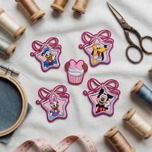Op de afbeelding: Geborduurde patches met Disney-personages Donald Duck, Pluto, Daisy Duck en Mickey Mouse, plus een cupcake. Elke patch is omlijst door een roze ster en strik. Naaibenodigdheden, waaronder garenklosjes, een schaar en een meetlint, zijn ook zichtbaar.