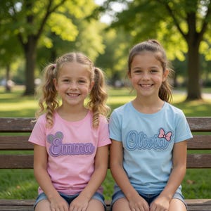 May include: Two young girls wearing matching t-shirts with personalized names. One shirt is pink with "Emma" in a purple font and a mouse-ear design. The other is light blue with "Olivia" and a pink bow.