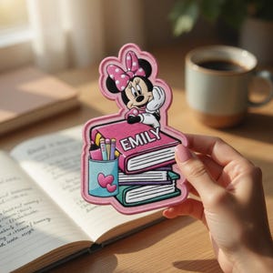May include: A pink embroidered patch featuring Minnie Mouse sitting on a stack of books. The top book has the name "EMILY" on it. A pencil holder with pencils and hearts is also on the patch. The patch is held in a person's hand.