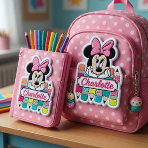 Op de afbeelding: Roze rugzak en etui set met witte stippen. Beide items hebben een Minnie Mouse patch met de naam "Charlotte". De etui bevat kleurrijke potloden. Een schattige schoolset voor kinderen.