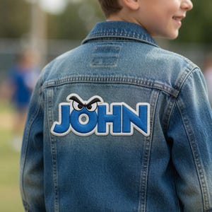 以下が含まれることがあります： 青いデニムジャケット。青い文字で「JOHN」と書かれた白い縁取りのパッチが付いています。パッチには名前の上に漫画の目のデザインがあります。ジャケットは人が着ています。