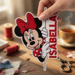 May include: A fabric embroidered patch featuring a cartoon mouse with a large red bow and a red and white polka dot dress. The mouse is holding a vertical white sign with the name ISABELLA printed in bold red capital letters.