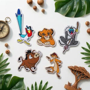 Puede incluir: Una colección de cinco parches bordados con personajes animados: un pájaro azul y rojo, un león joven, un mono gris con un bastón, un jabalí marrón y una suricata beige, dispuestos sobre una superficie blanca con elementos decorativos.