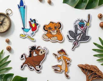 May include: A collection of five embroidered patches featuring animated characters: a blue and red bird, a young lion, a grey monkey holding a staff, a brown warthog, and a tan meerkat, all arranged on a white surface with decorative items.