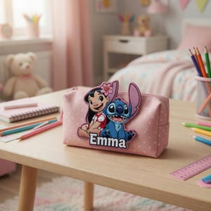 May include: A light pink pencil case with a white polka dot pattern featuring a raised applique of Lilo and Stitch with the name Emma printed in white letters. The rectangular case has a zipper closure and sits on a wooden desk surface.