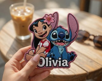 May include: An embroidered patch featuring the characters Lilo and Stitch. Lilo has black hair with a pink flower, wearing a red and white patterned top. Stitch is blue with large ears. The name Olivia is embroidered in white at the bottom.