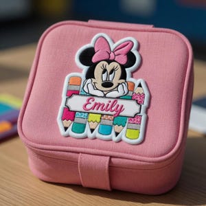 Op de afbeelding: Een roze, vierkante sieradendoos met een Minnie Mouse-applicatie. De applicatie toont Minnie Mouse met een roze strik, potloden en de naam "Emily". De doos heeft een sluiting.