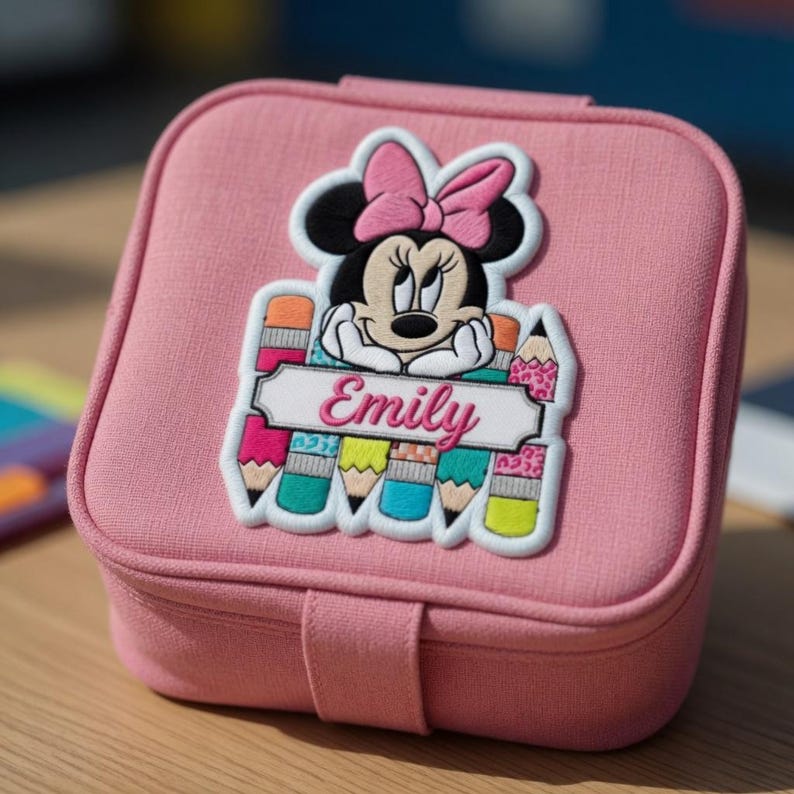 Op de afbeelding: Een roze, vierkante sieradendoos met een Minnie Mouse-applicatie. De applicatie toont Minnie Mouse met een roze strik, potloden en de naam "Emily". De doos heeft een sluiting.