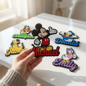 May include: Five embroidered character patches featuring Mickey Mouse with the name Michael, Goofy with Christopher, Pluto with James, Donald Duck with Donald, and Daisy Duck with Emily. Each patch displays a colorful cartoon figure above a name.