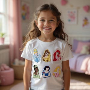May include: A white t-shirt featuring colorful patches of Disney princesses. The patches include images of Cinderella, Belle, Ariel, Tiana, Snow White, and Aurora. The shirt is worn by a young girl with long brown hair.