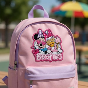 May include: Pink backpack with a purple handle and accents. The front features a colorful patch with Minnie Mouse and Daisy Duck wearing sunglasses and holding ice cream. The word "Besties" is below the characters.