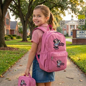 Puede incluir: Una mochila rosa y una bolsa de almuerzo a juego con un dise&ntilde;o de Minnie Mouse. La mochila tiene un bolsillo frontal y el nombre "Emily" bordado. La ni&ntilde;a lleva una camisa a cuadros rojos y blancos y una falda vaquera.