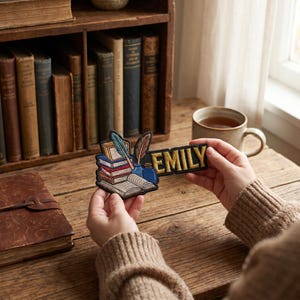 Peut inclure: Écusson avec le nom "EMILY" en lettres dorées, devant une pile de livres, une plume et un encrier. L'écusson est posé sur un bureau en bois, avec des livres et une tasse de thé en arrière-plan.