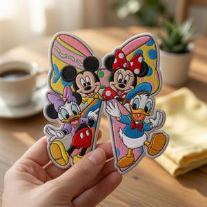 May include: An embroidered patch shaped like a bow featuring Mickey Mouse, Minnie Mouse, Daisy Duck, and Donald Duck. The background of the bow has a colorful pattern with pink, yellow, and blue sections. The characters are depicted in their classic colors.