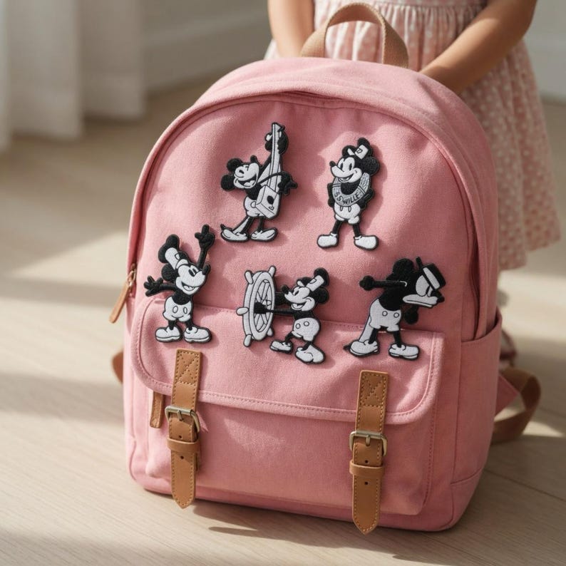 Op de afbeelding: Een roze rugzak met bruine leren riemen en gespen. De rugzak is versierd met verschillende zwart-witte cartoonkarakterpatches. Hij heeft een voorvak en een handvat aan de bovenkant.