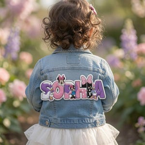 May include: A light blue denim jacket featuring a large, custom embroidered patch on the back. The patch displays the name SOPHIA in purple and pink lettering, decorated with various Minnie Mouse and Daisy Duck character illustrations.