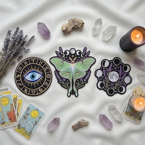 Puede incluir: Un surtido de objetos místicos dispuestos sobre una tela blanca. Incluye cartas del tarot, parches con diseños celestiales, cristales, una vela encendida y lavanda seca. Los parches presentan un ojo, una polilla y un diseño de fases lunares.