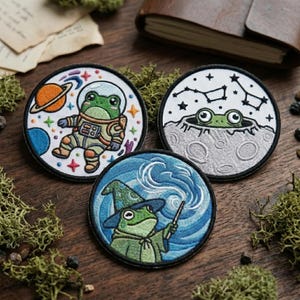 May include: Three embroidered patches featuring whimsical frog designs. One shows an astronaut frog, another a frog peeking from a moon, and the last a wizard frog with a wand. Each patch is circular with a black border, set against a wooden surface.